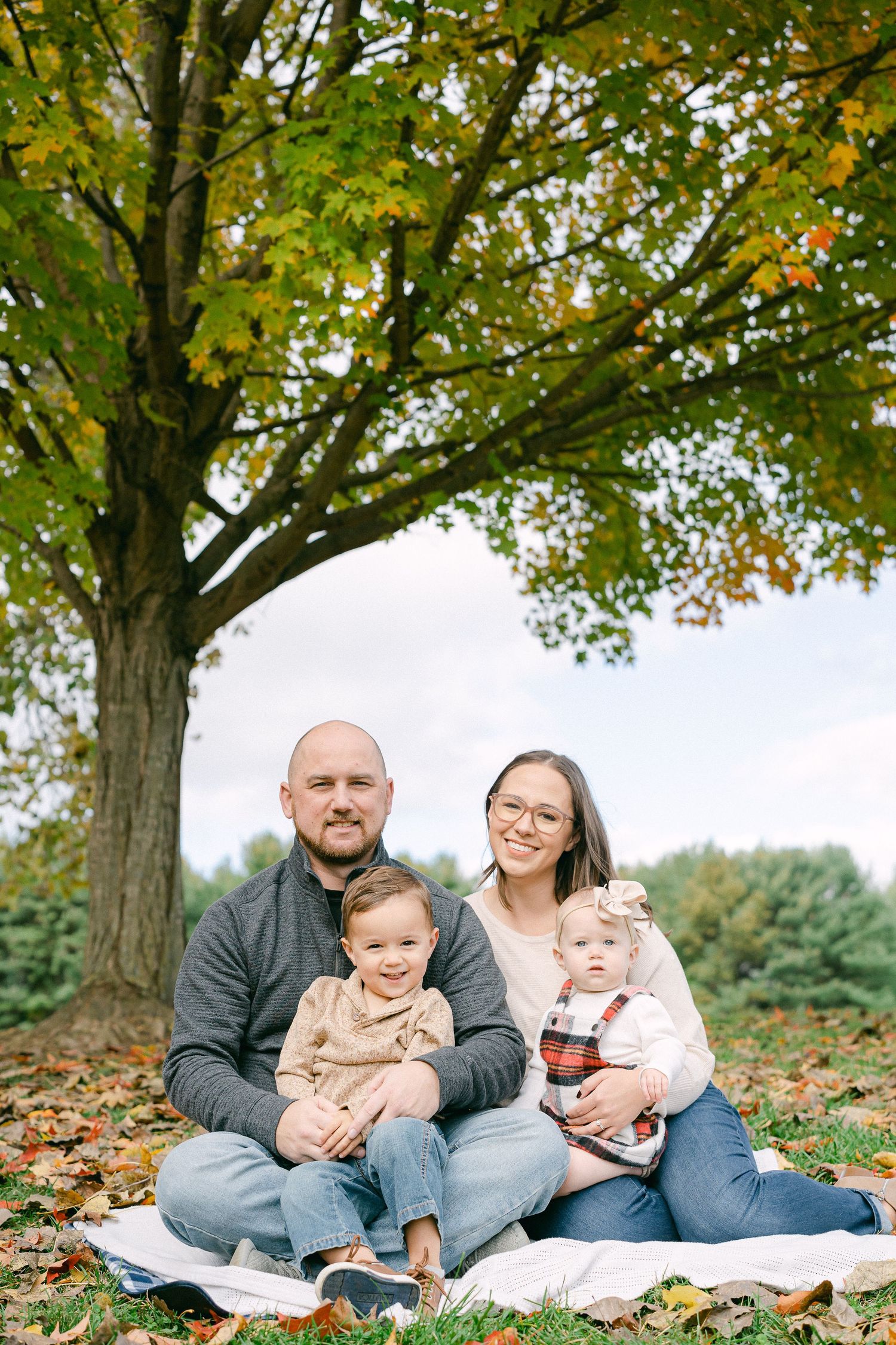 A family sits together on a white blanket surrounded by autumn leaves under mature trees.