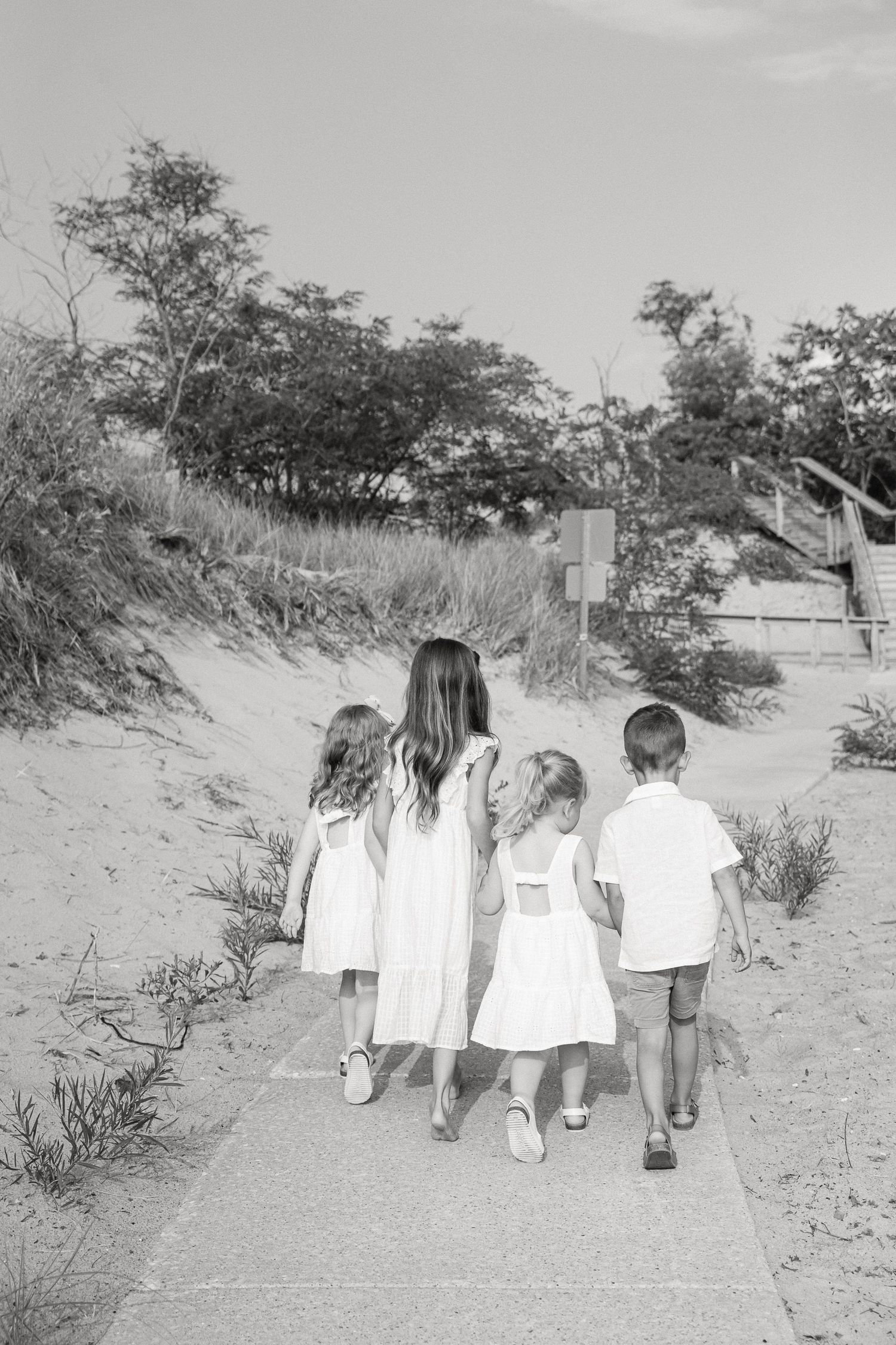 A group of people walk together along a sandy beach path through coastal vegetation in black and white.