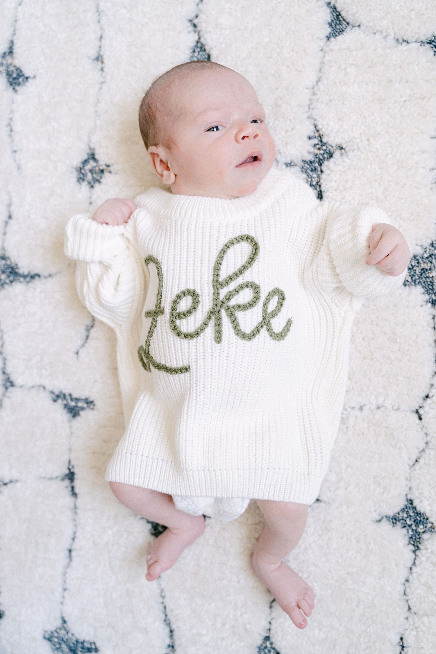 Baby lying on white patterned rug wearing cream colored sweater with text.