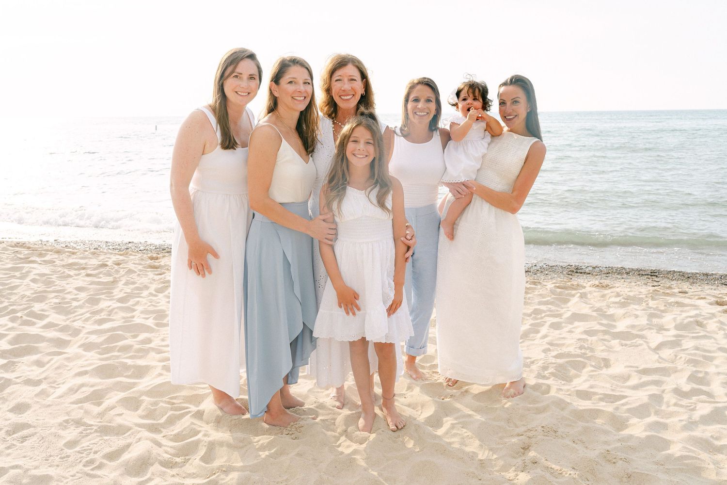 A group of people in white and blue dresses pose together on a sunny beach by the ocean.