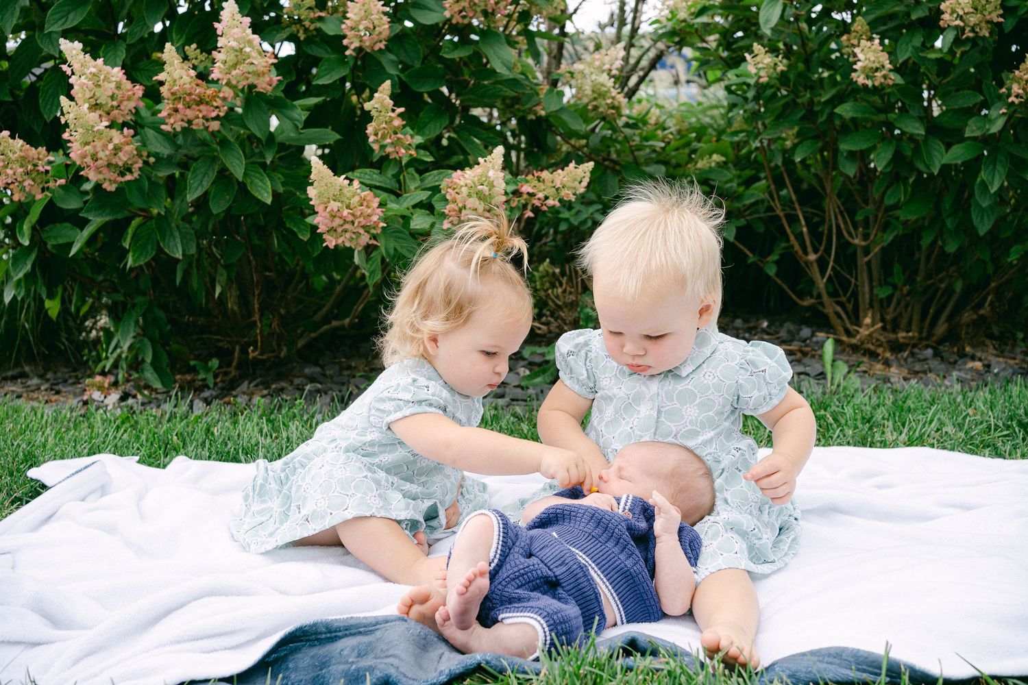 Two young children sit together on a white blanket in a garden setting with flowering bushes in the background.