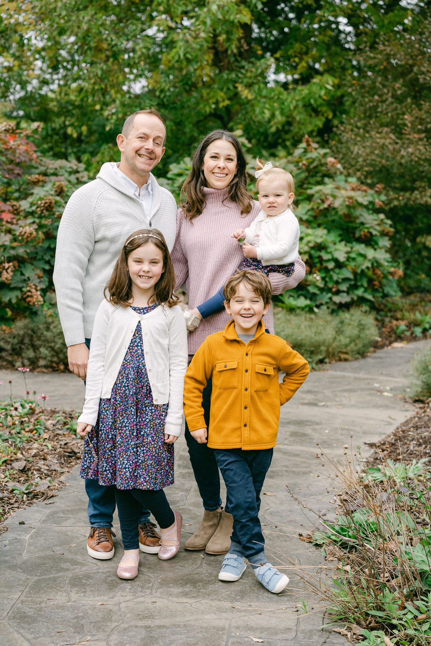 Family wearing coordinated casual outfits poses together on stone path surrounded by lush greenery in outdoor setting.