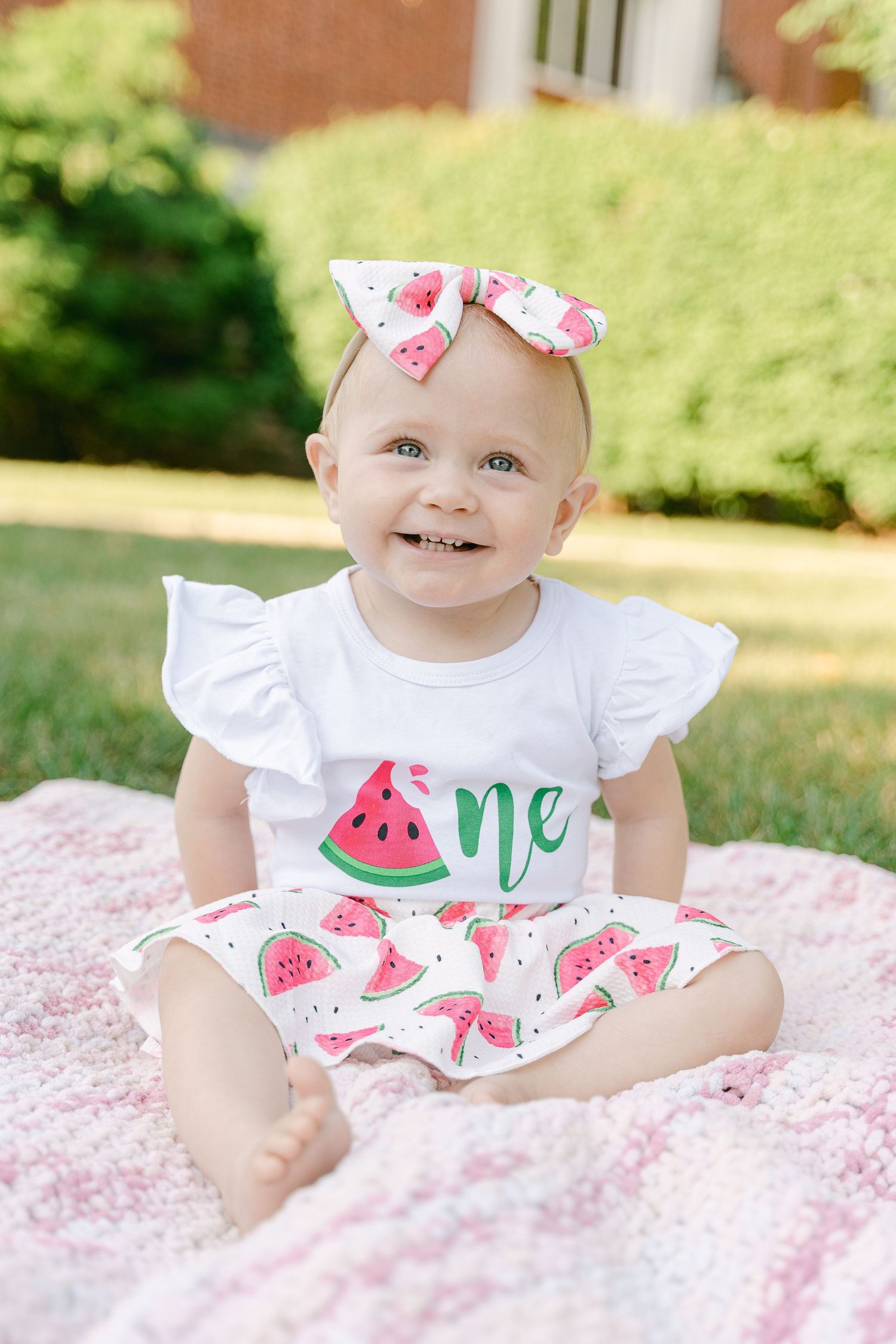 Baby wearing white watermelon-themed outfit lies on pink blanket during outdoor summer photoshoot.