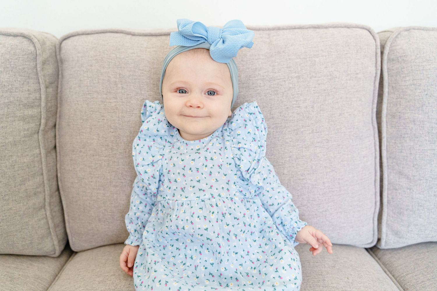 Baby in light blue floral dress with matching bow headband lying on gray couch.