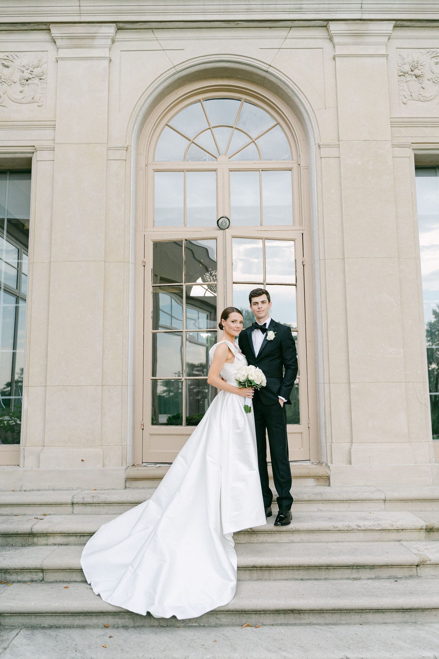 Bride and groom posing on stone steps before an elegant arched window at their wedding venue in Philadelphia, PA