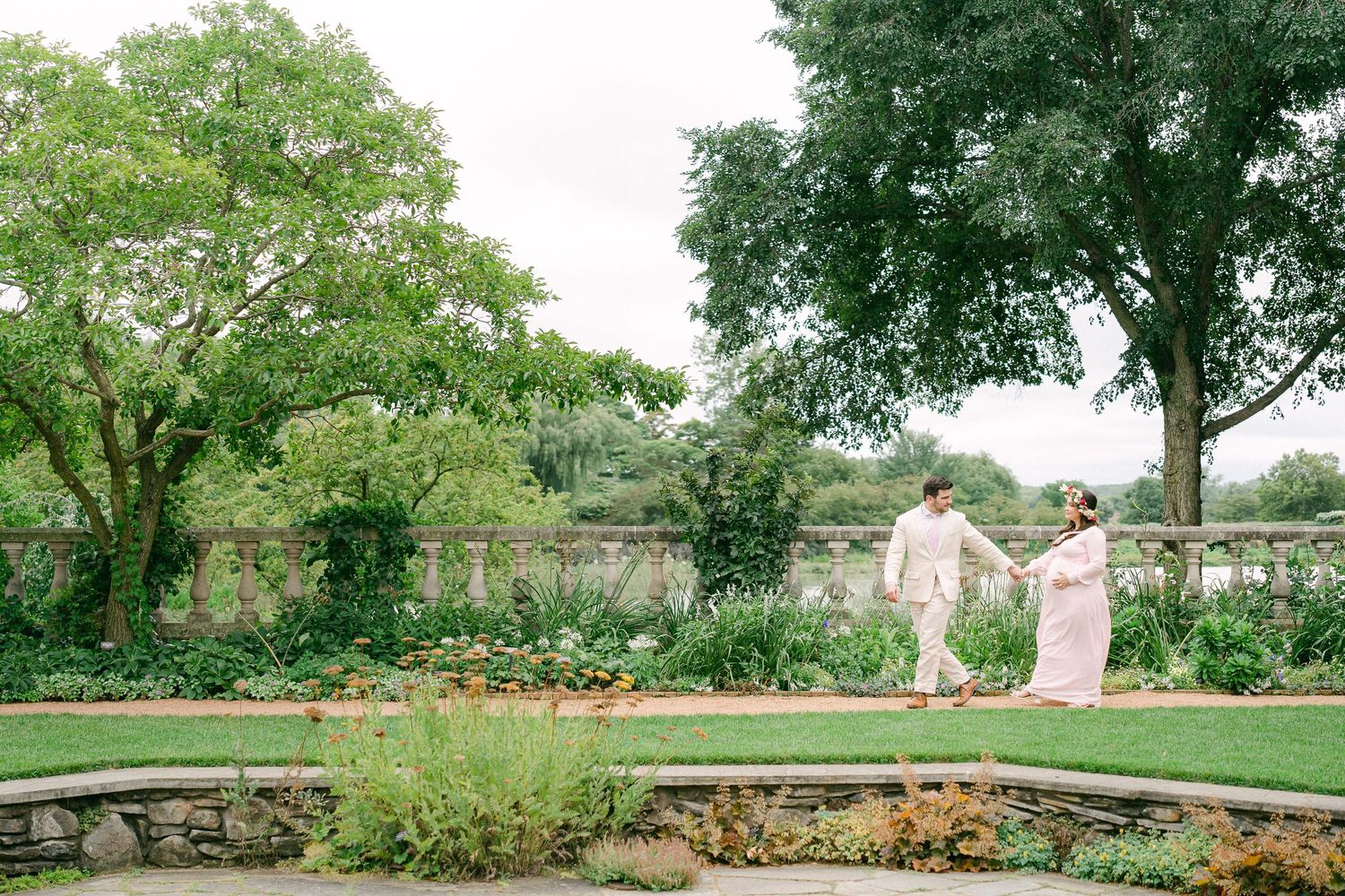 Couple walks through lush garden pathway surrounded by blooming flowers and mature trees in summer setting.