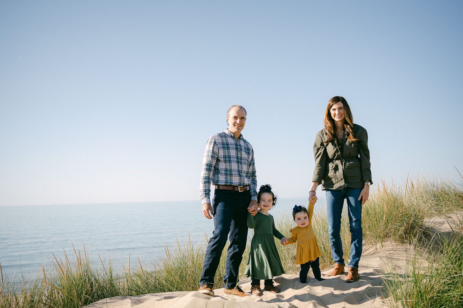 Family walking together on sandy beach dunes with tall grass and ocean view during sunset.