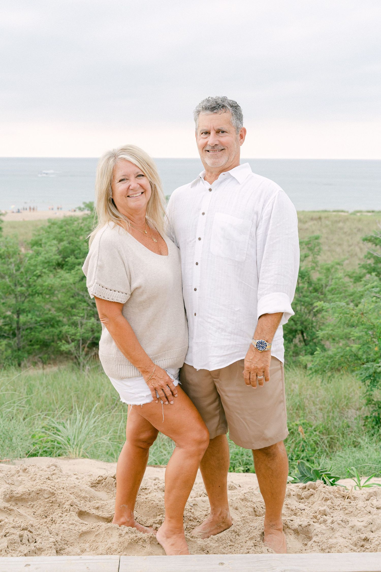 A couple in white and tan outfits poses playfully together on a sandy beach with green foliage in the background.