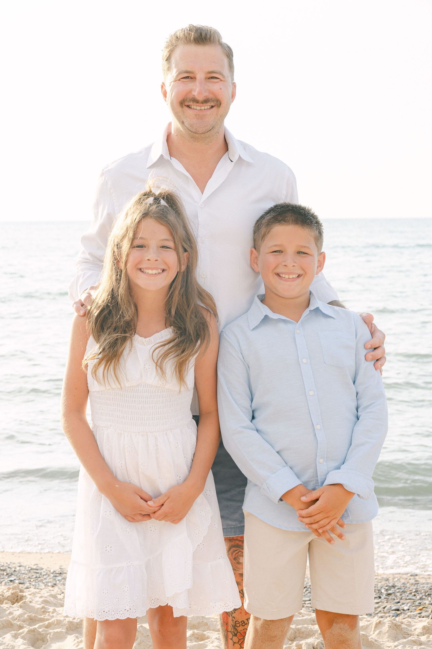 Family portrait at the beach with three people wearing light colored clothing standing together by the ocean.