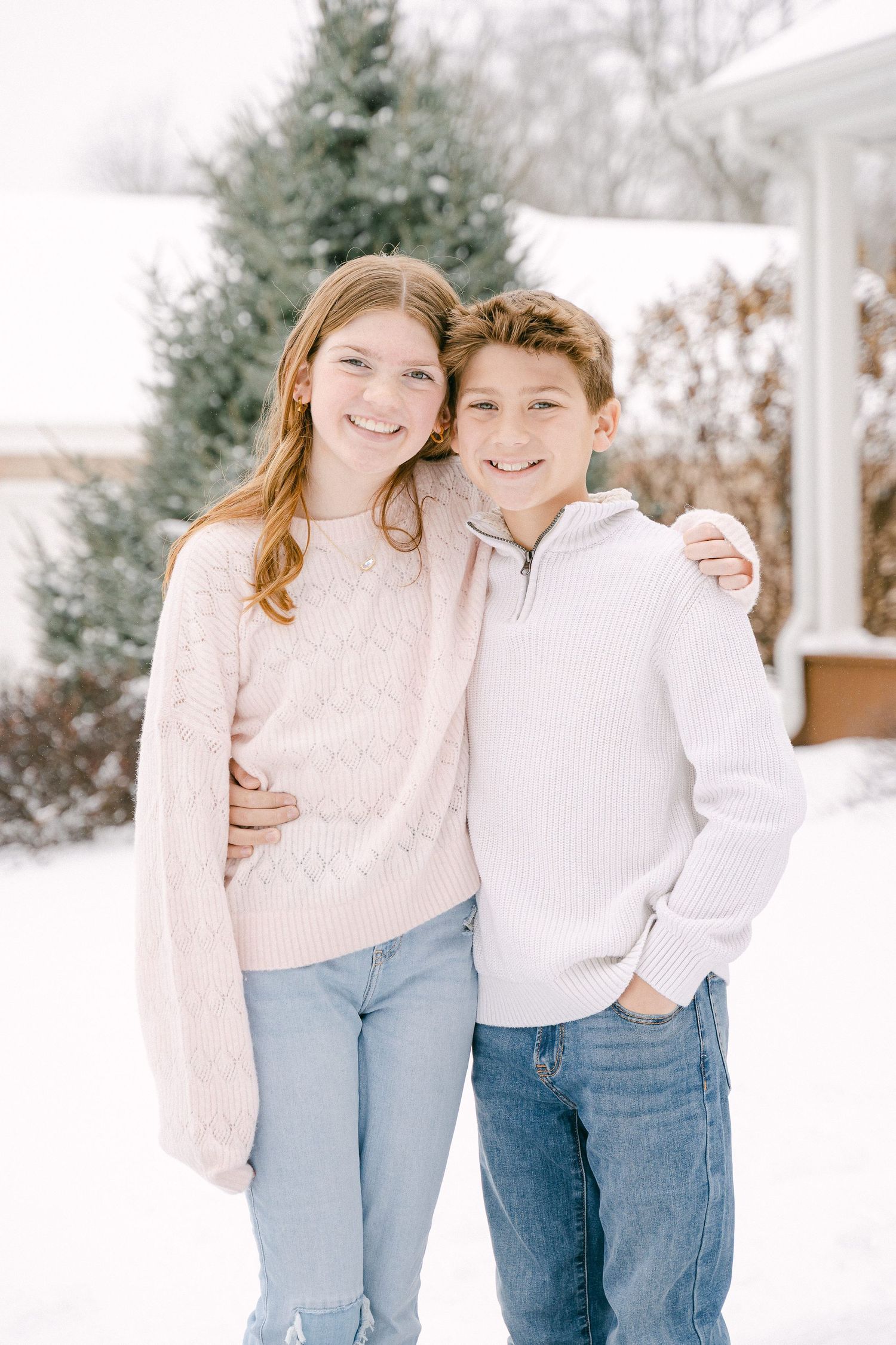 Two siblings pose together in winter sweaters against a snowy outdoor backdrop with evergreen trees.