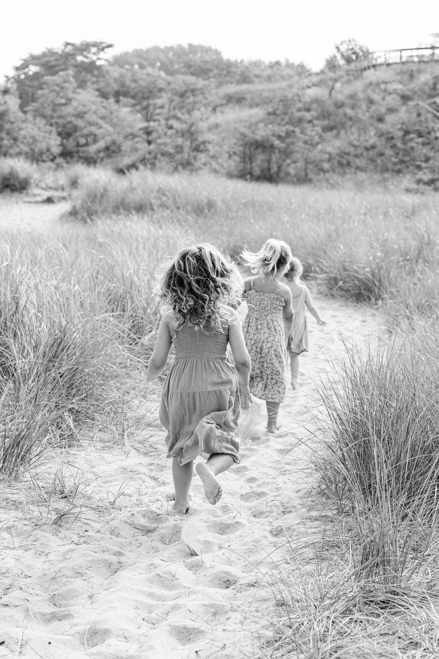 Two figures in flowing dresses walk along a sandy beach path through tall grass on a sunny summer day.