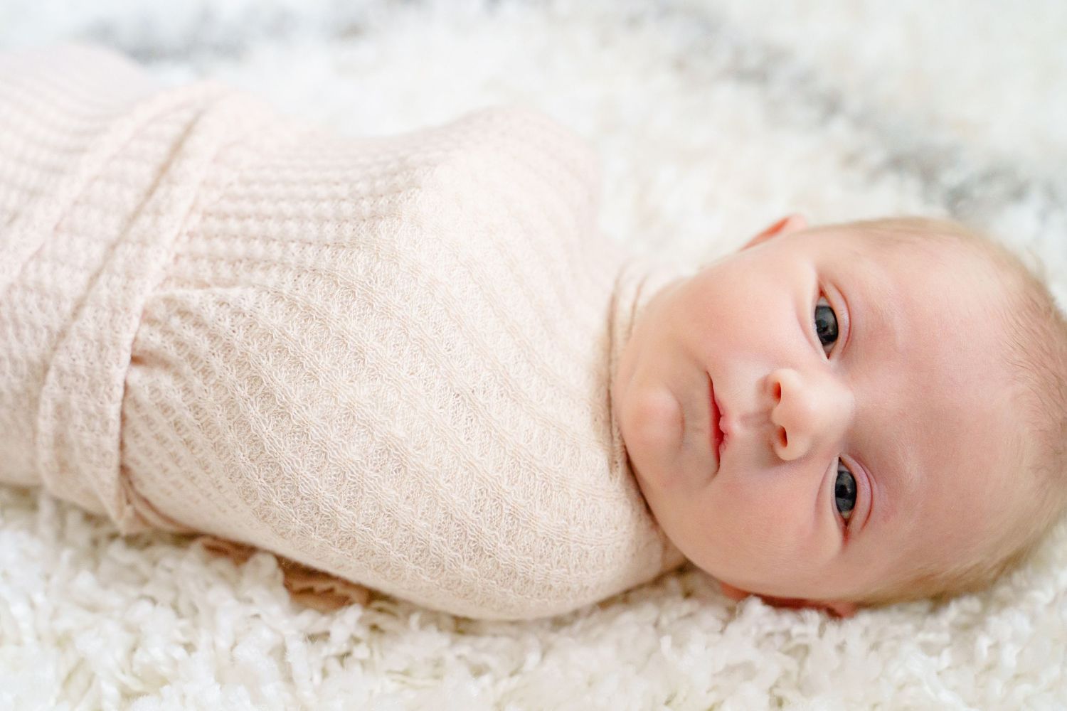 A baby wearing a cream colored sweater lies on white bedding while looking at the camera.