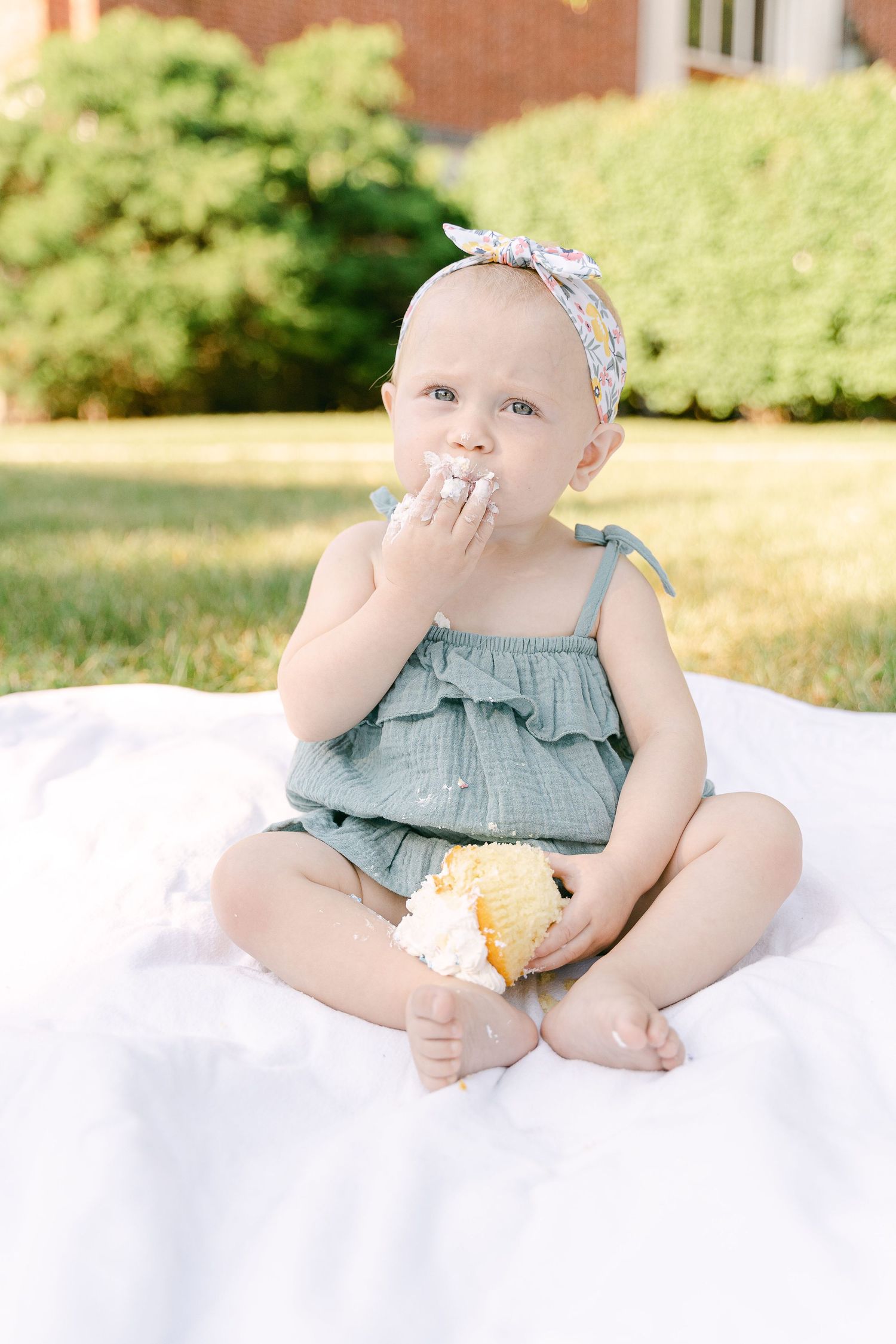 A small child in a ruffled blue dress sits on a white blanket outdoors enjoying a snack.