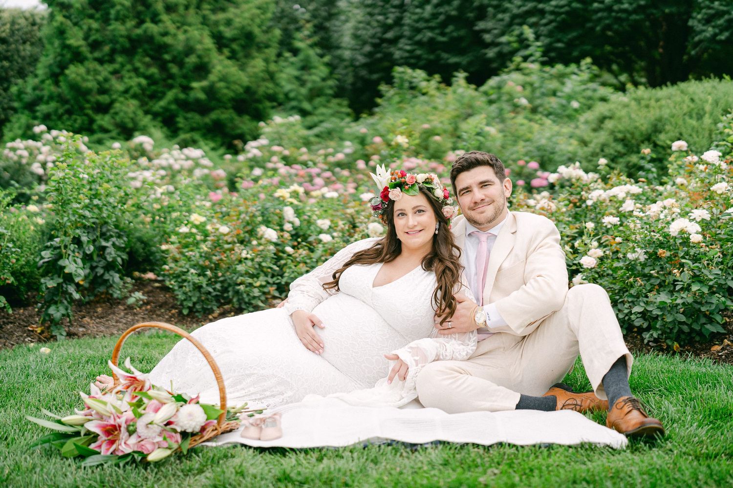 A couple in white outfits lounges on a blanket in a lush garden surrounded by blooming flowers and greenery.