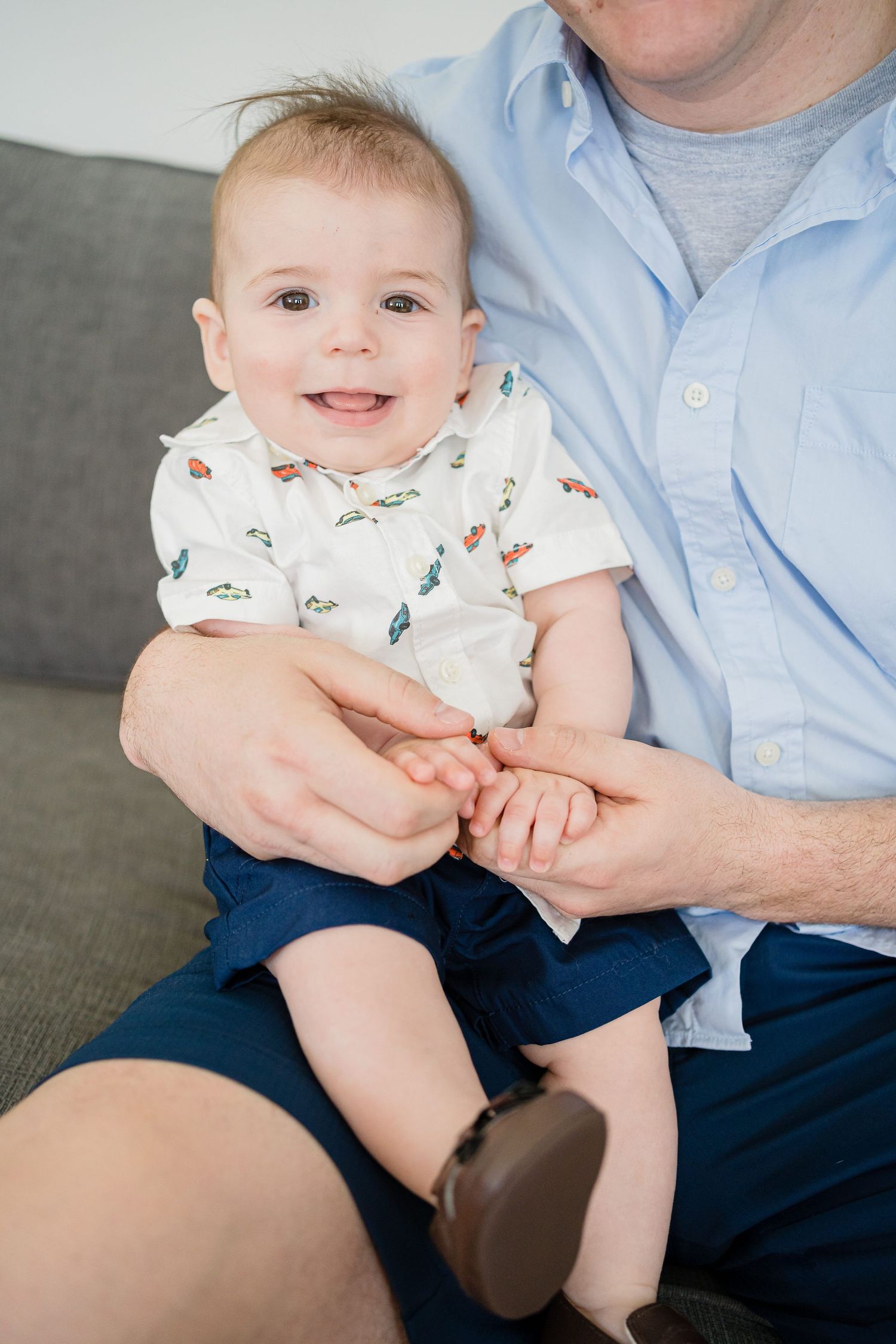 Baby in white patterned outfit sits on lap wearing navy blue shorts.
