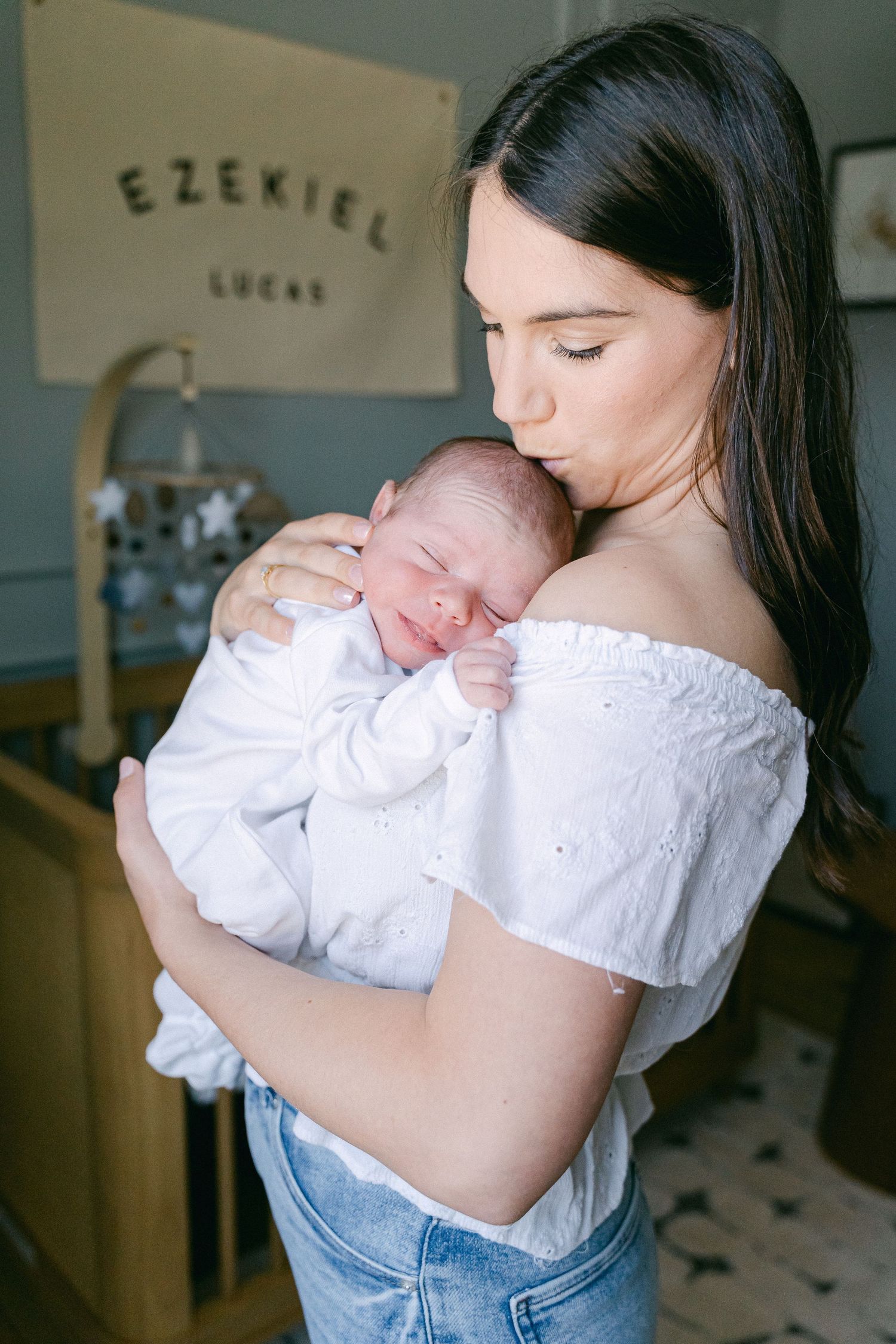 Mother in white top and jeans holds sleeping newborn baby in tender embrace.