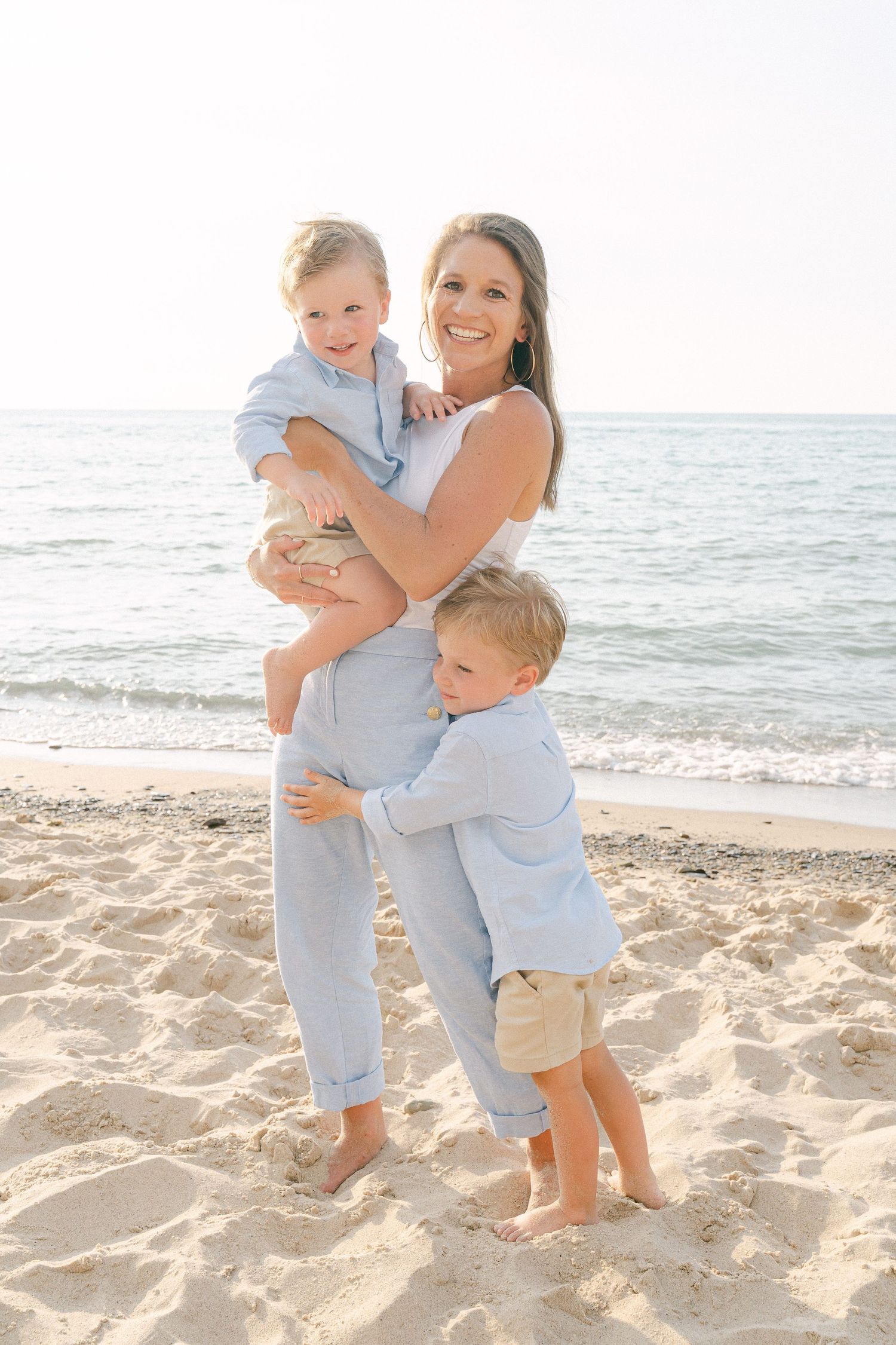 Family wearing coordinated light blue and white outfits embracing together on a sunny beach at sunset.