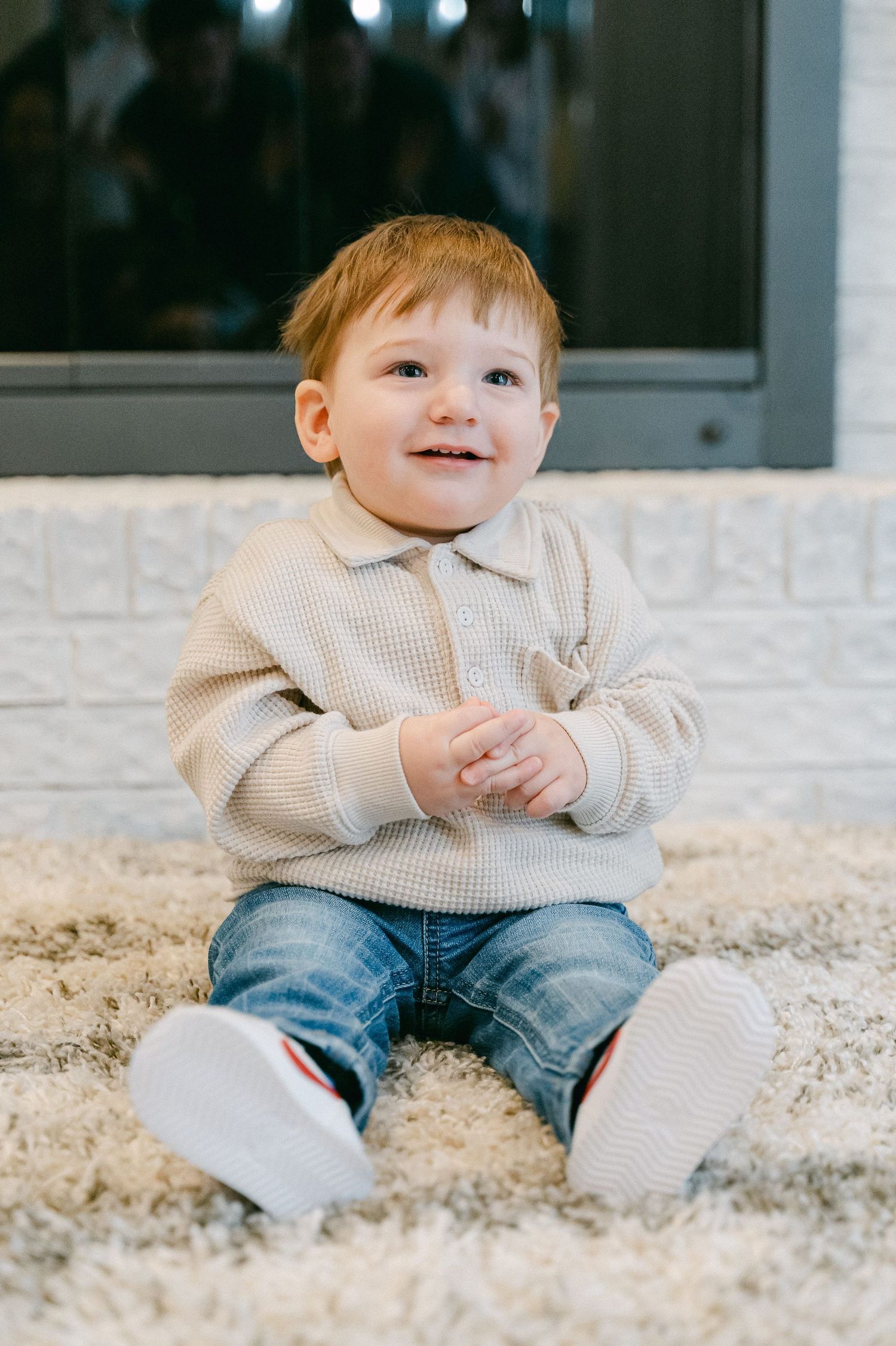 Small child in beige sweater and jeans sitting on plush carpet with a happy expression.