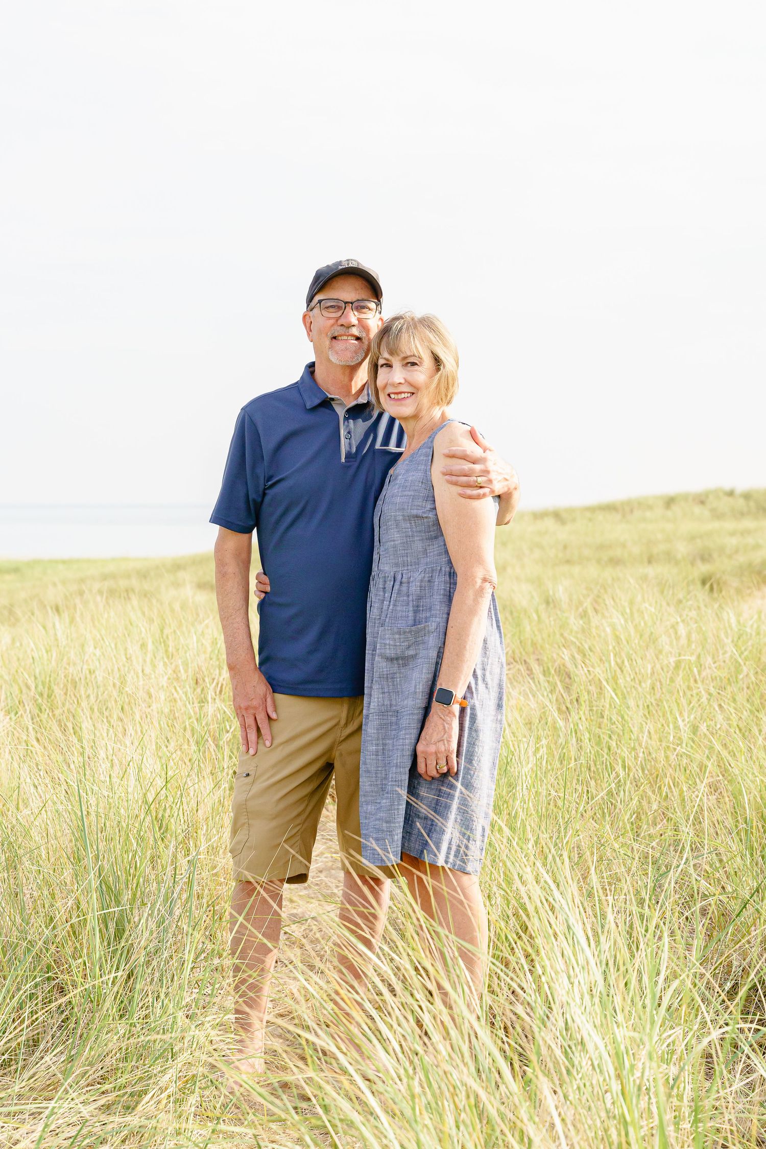 A couple embraces while standing in a sunlit field of tall grass during a summer photoshoot.