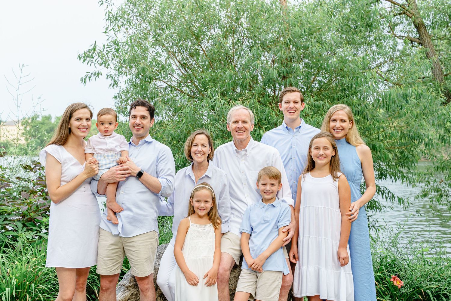 A family group poses together outdoors in coordinated light blue and white outfits near a lake on a sunny day.