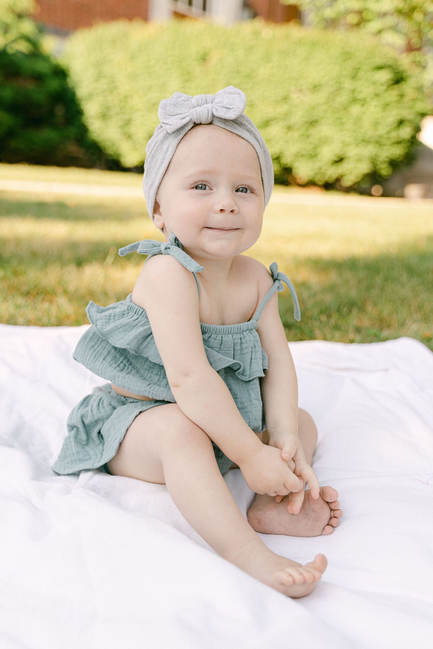 Baby in mint green romper and headband lying on white blanket outdoors.
