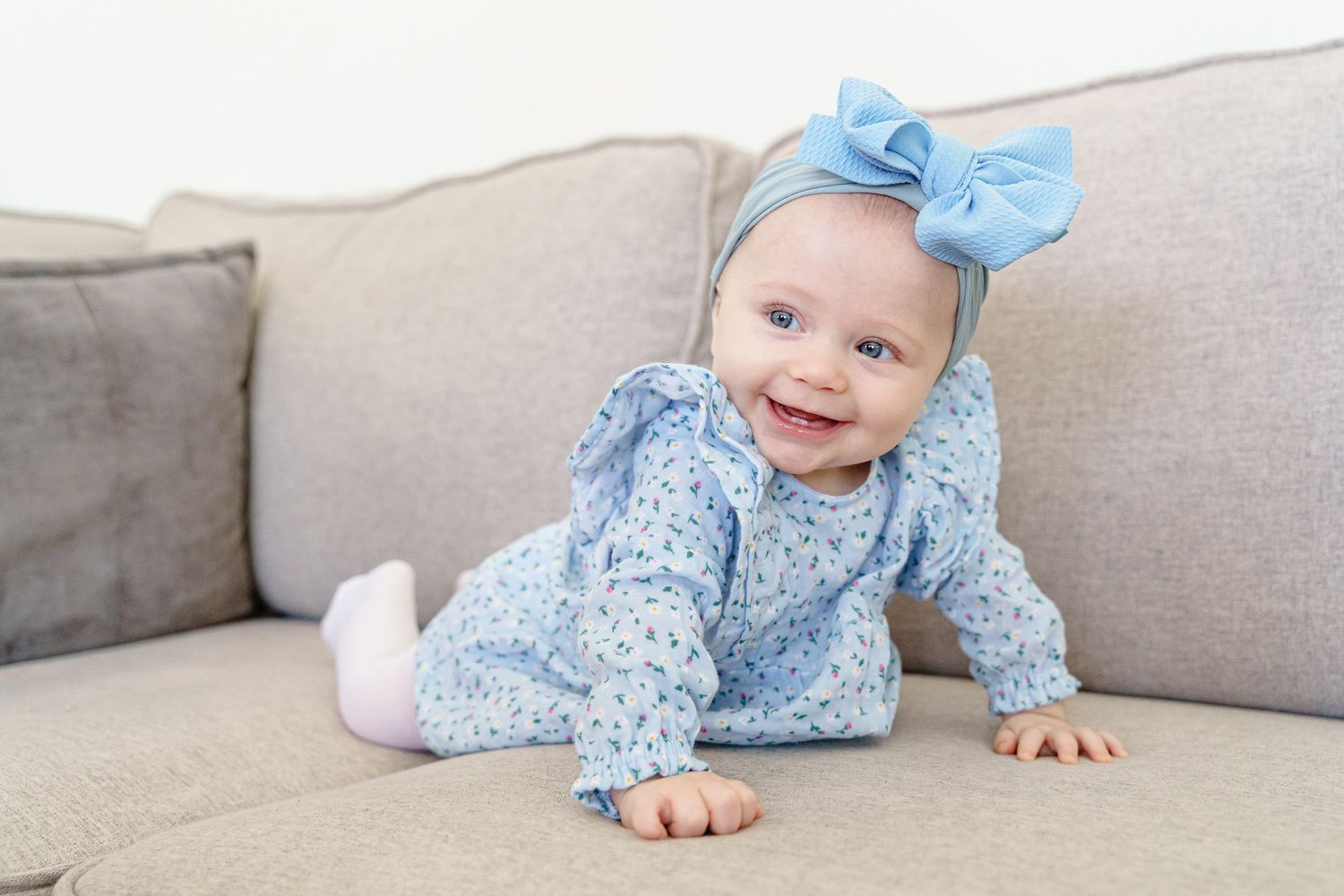 Baby wearing blue floral dress with bow crawling on gray sofa.