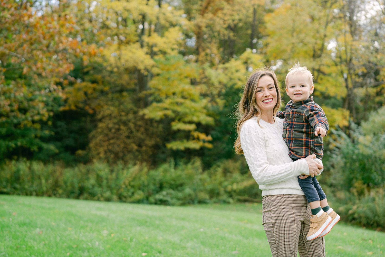 A parent and child stand together on a grassy lawn with autumn foliage in the background.