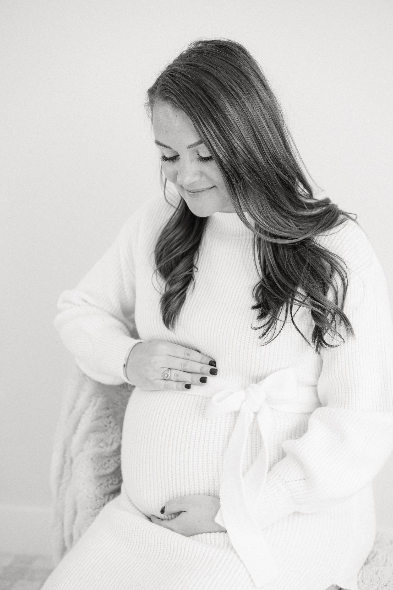 Expectant mother in white sweater dress gently touches her baby bump.