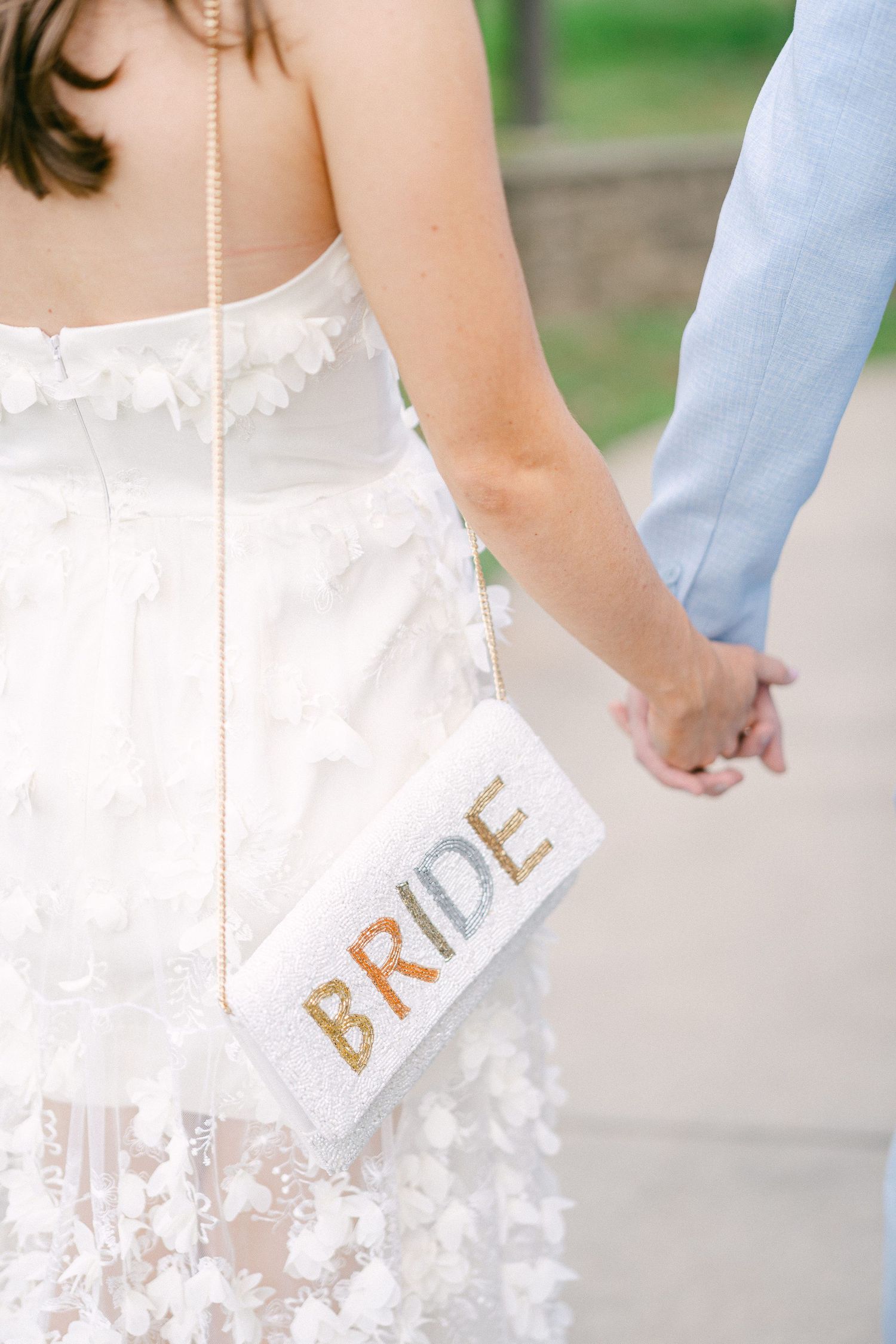 Bride holding hands with groom, wearing white floral dress and beaded BRIDE clutch bag at wedding in Philadelphia, PA