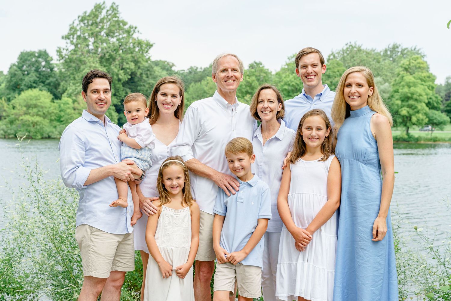 A family wearing coordinated blue and white outfits poses together outdoors near a lake on a sunny summer day.