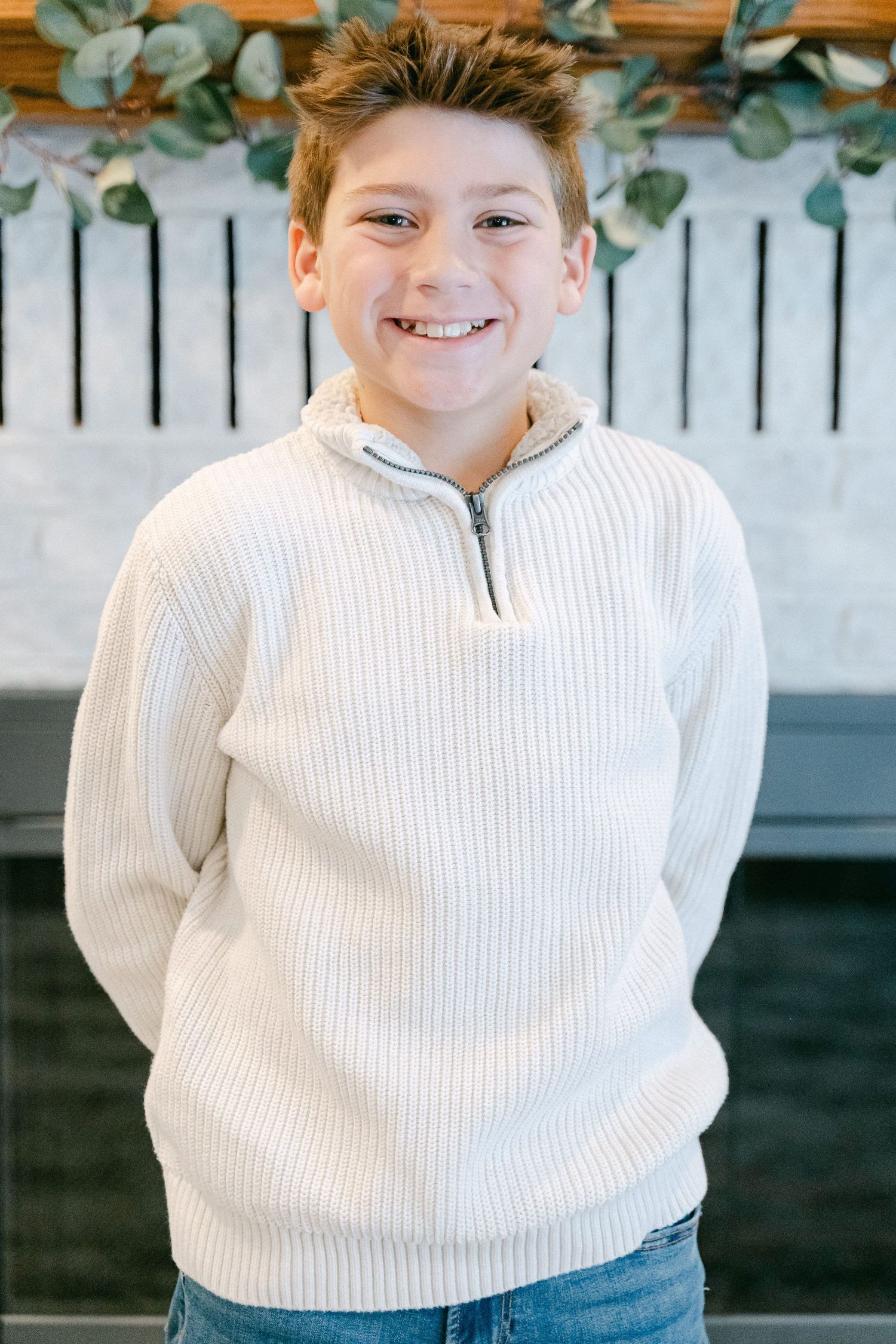 A young person in a white knit sweater smiles while standing against a white picket fence with greenery.