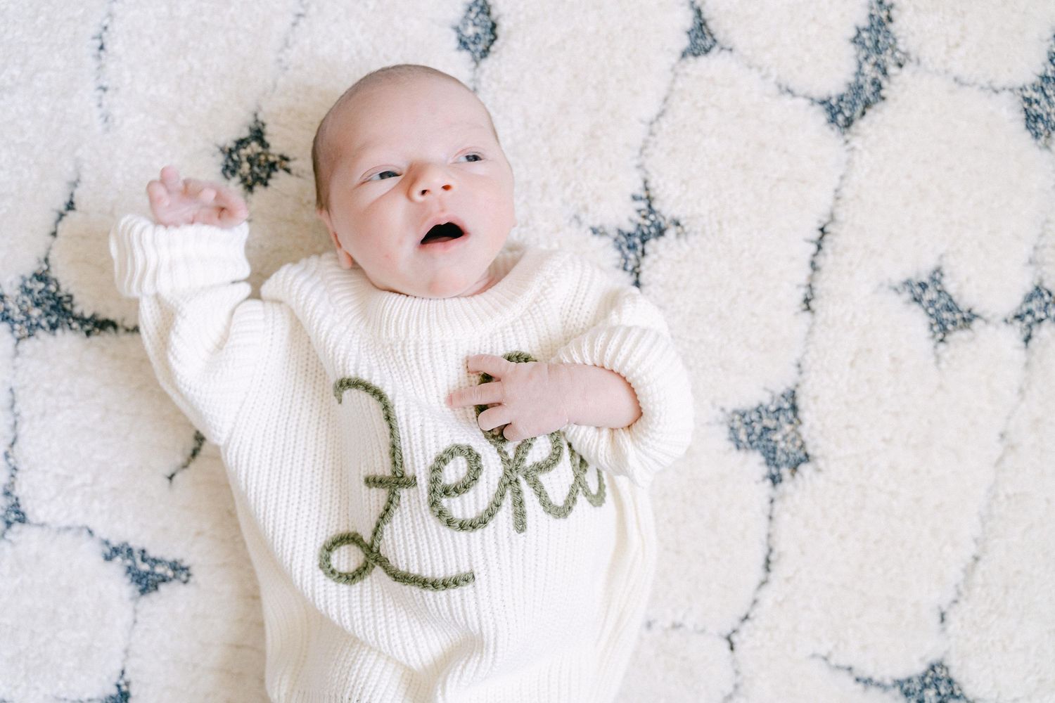 Baby wearing white knit clothing lies on a textured white blanket with delicate grey pattern.
