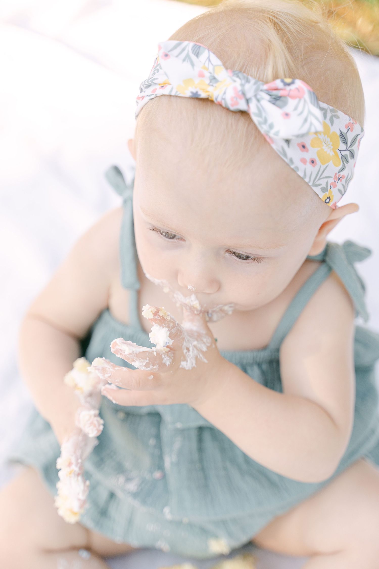 Baby in blue dress and floral headband enjoying cake during first birthday smash cake session.