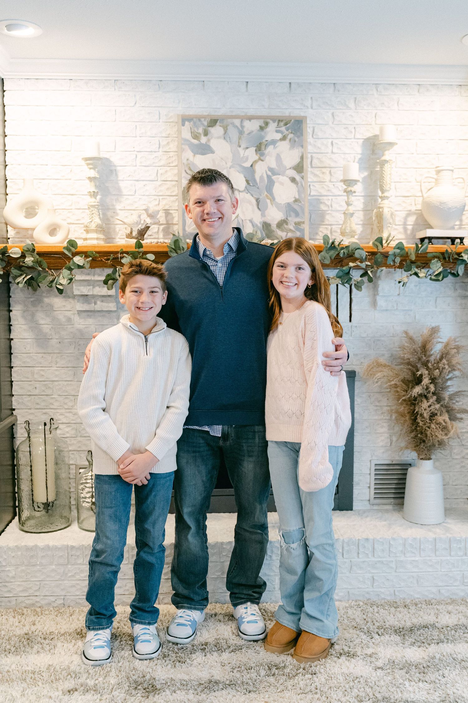 Three people stand together in front of a white brick fireplace decorated with holiday greenery and candles.