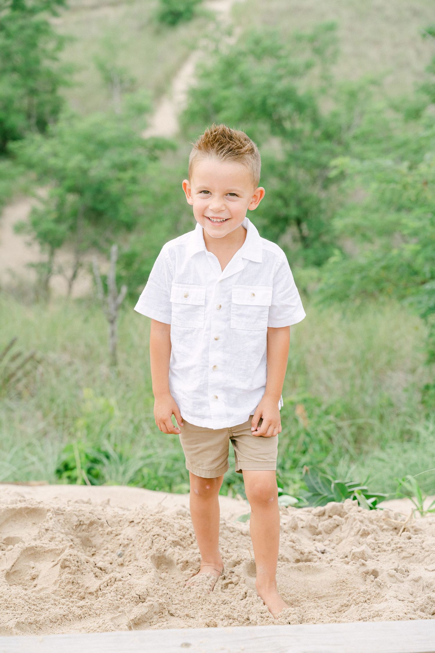 Child in white shirt and khaki shorts plays on sandy hill against green background.