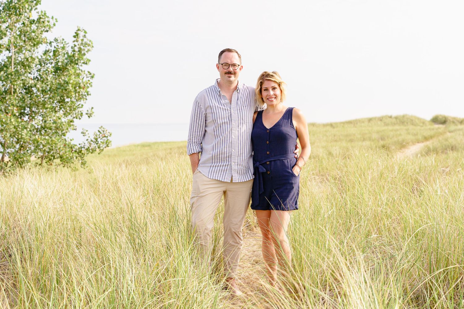 A couple stands together in a sun-drenched field of tall grass on a summer day.
