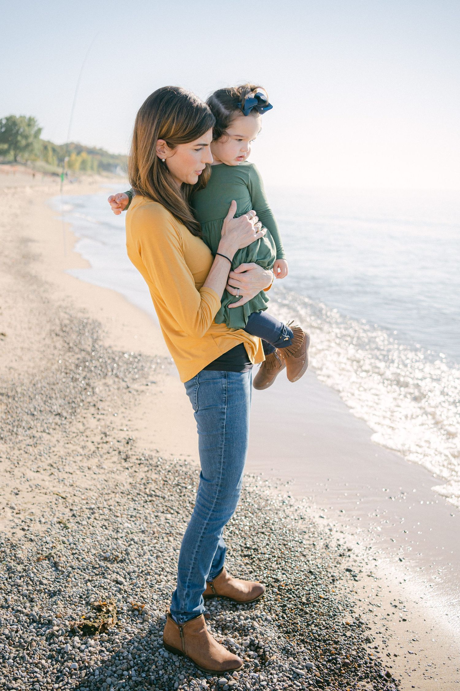 A parent in a yellow sweater holds a baby while standing on a rocky beach at sunset.