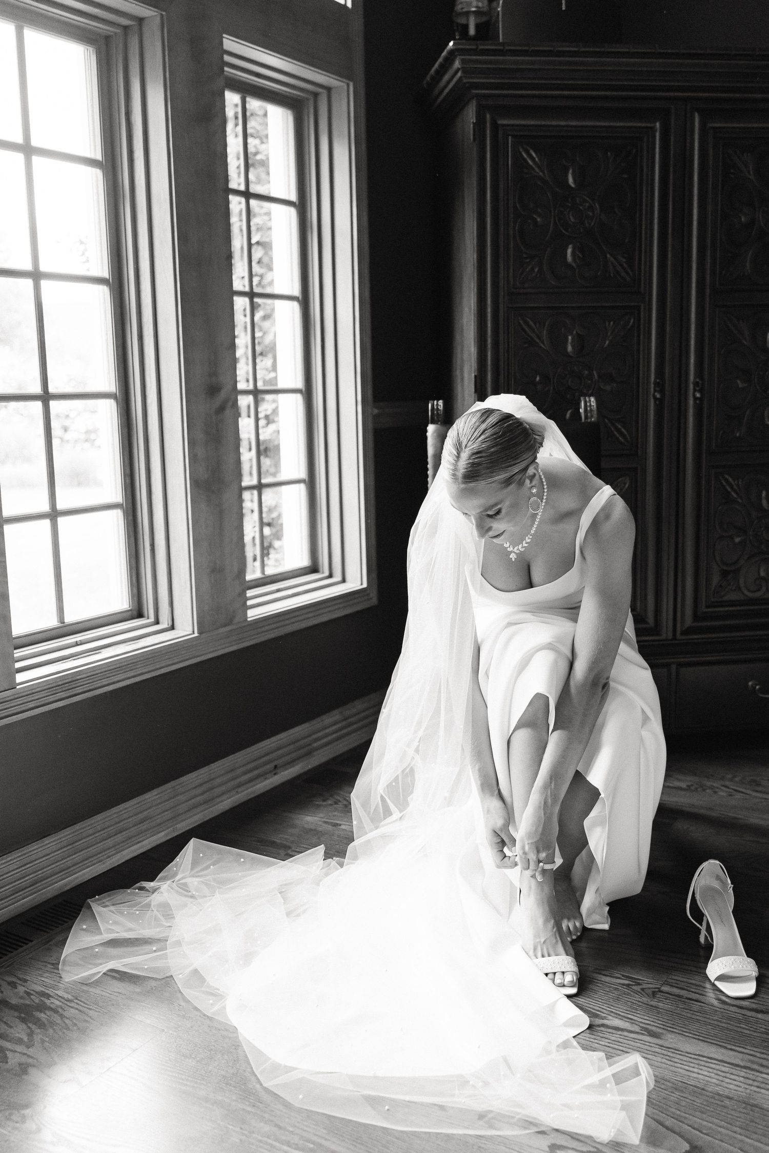 Bride in white gown and veil fastening her heels on hardwood floor near elegant windows in black and white photo in Valparaiso, IN