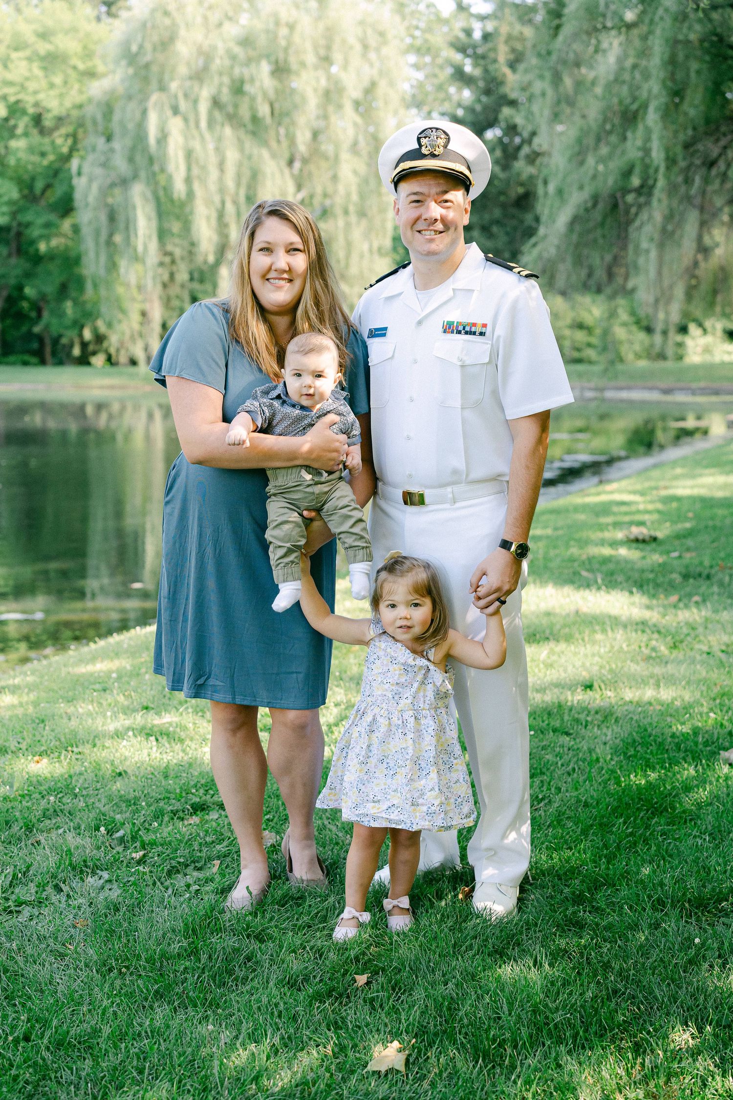A family poses together on a grassy lawn near a tranquil lake with willow trees in the background.