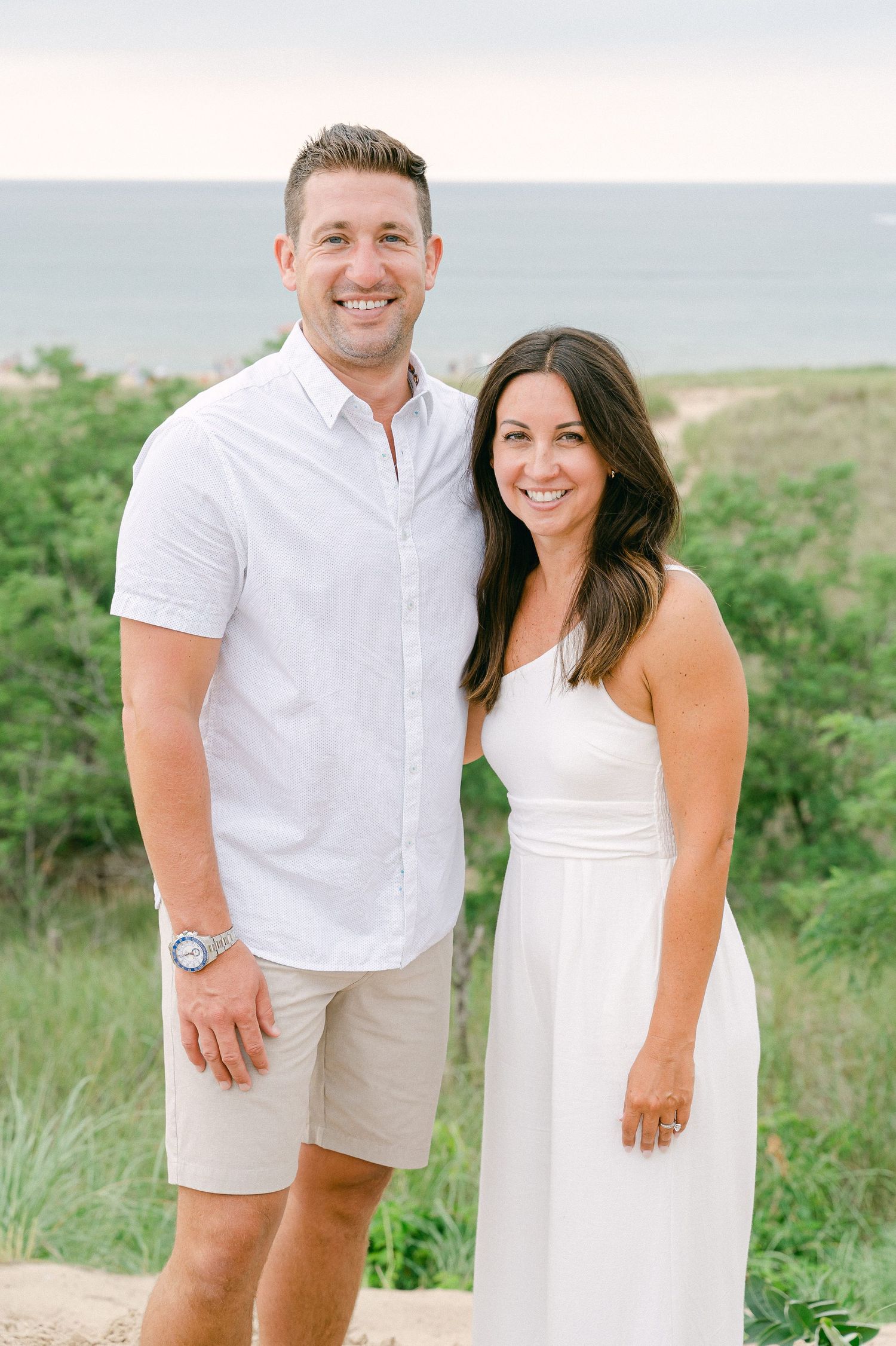 A couple in coordinating white outfits poses together on a grassy hillside during a casual outdoor photoshoot.