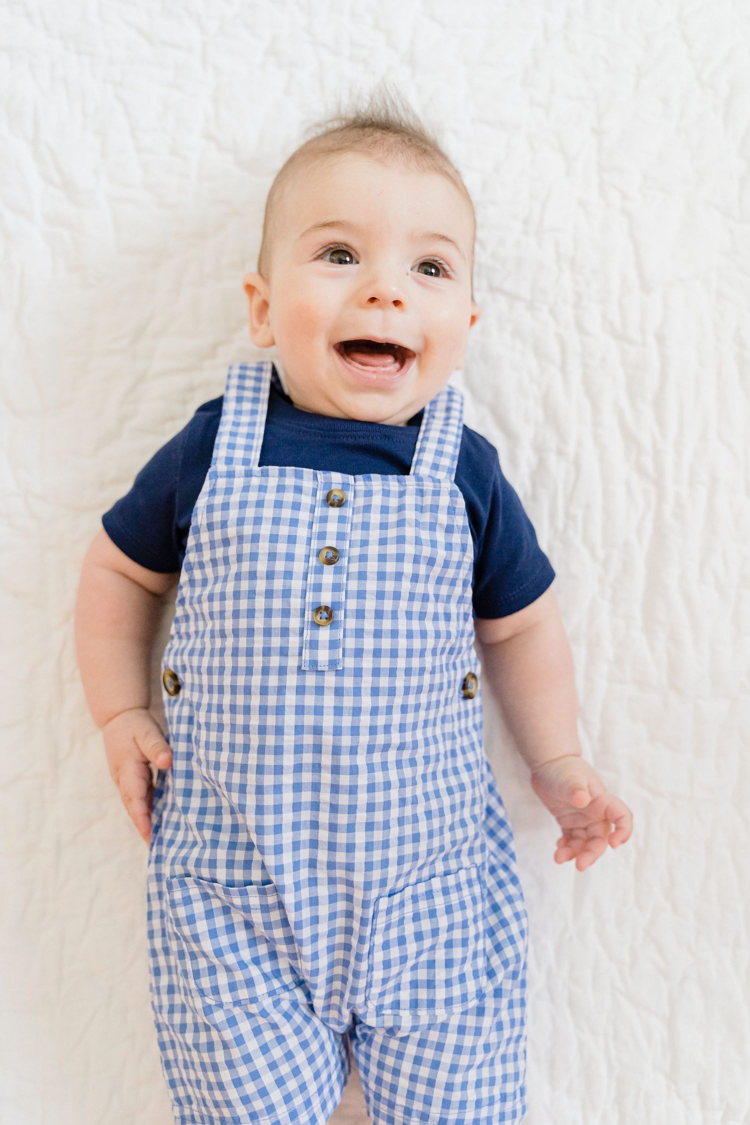 Baby wearing blue gingham overalls and navy shirt lies on white blanket with joyful expression.