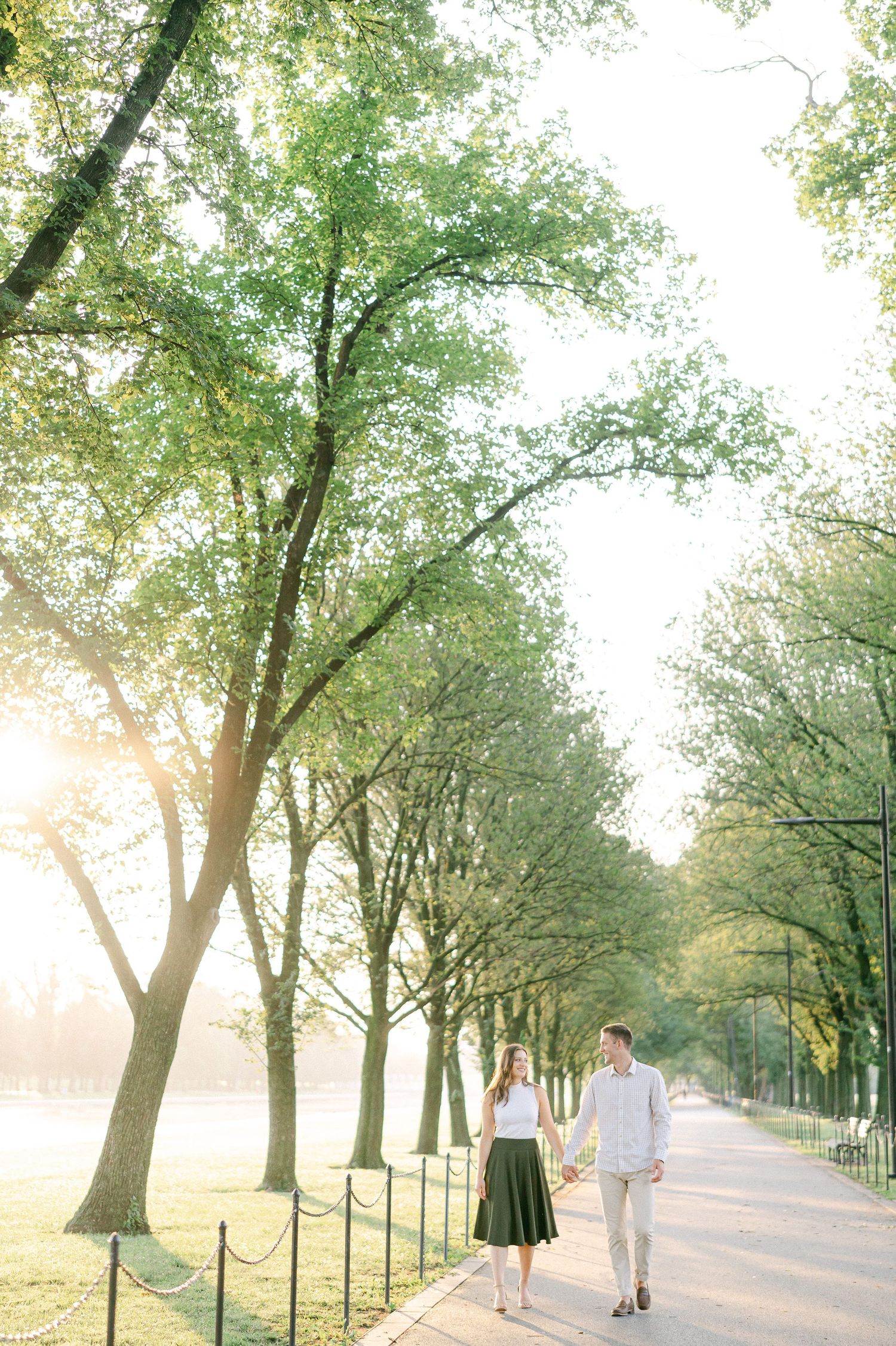 Couple walking together on a tree-lined path during golden hour with sunlight filtering through the leaves.