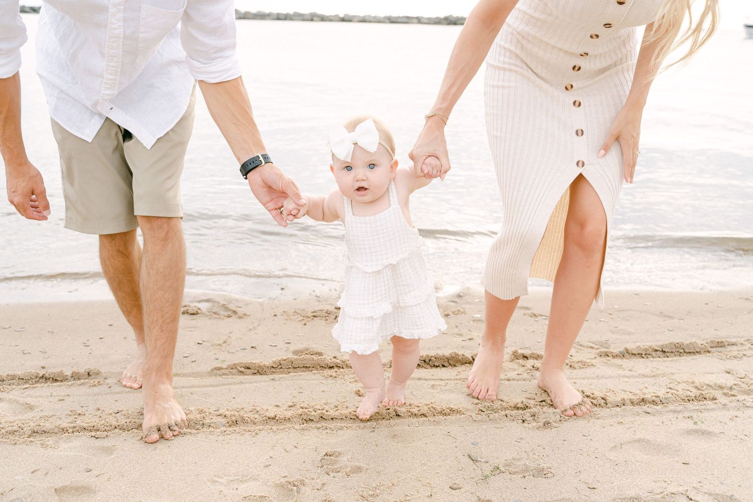 Parents in white clothing help a small child take first steps on a sandy beach.