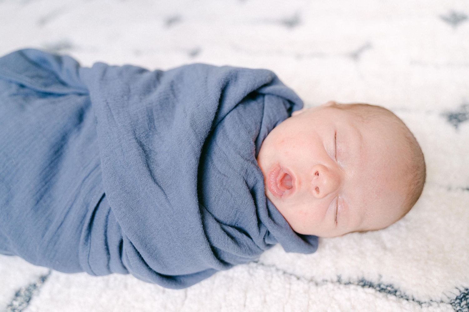 A sleeping newborn baby wrapped in soft blue swaddle blanket on white textured fabric.