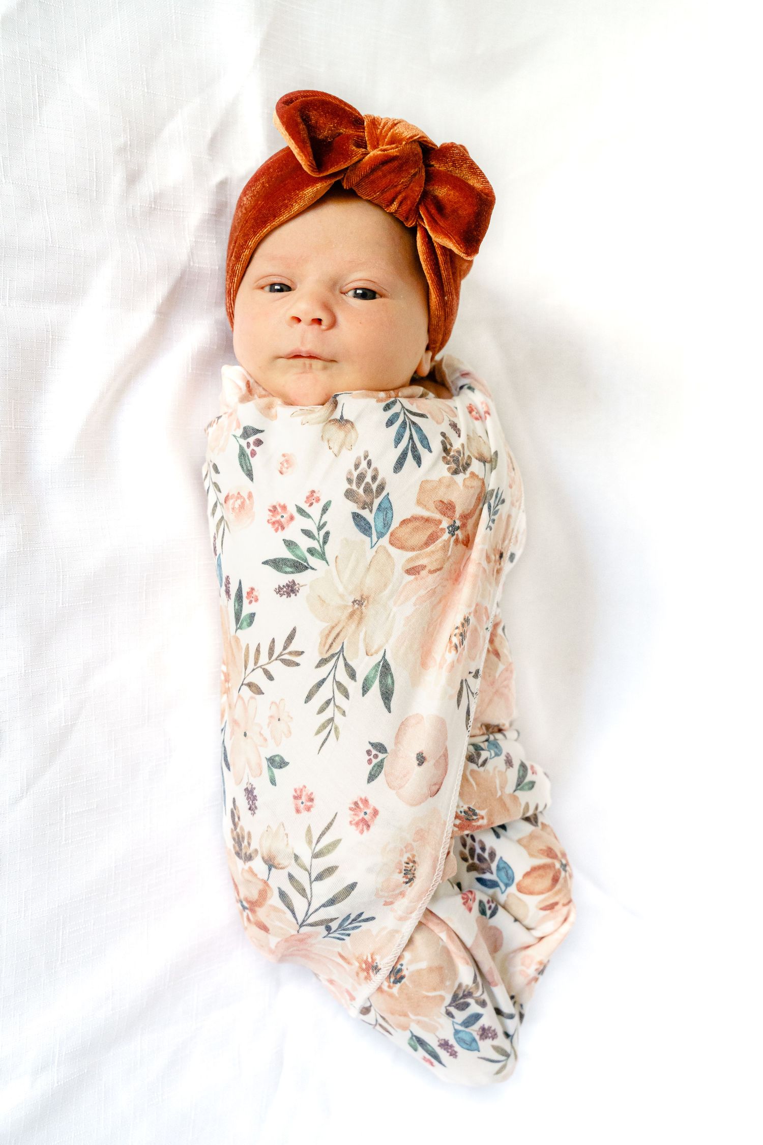 Baby wearing floral swaddle and orange bow headband lying on white background.