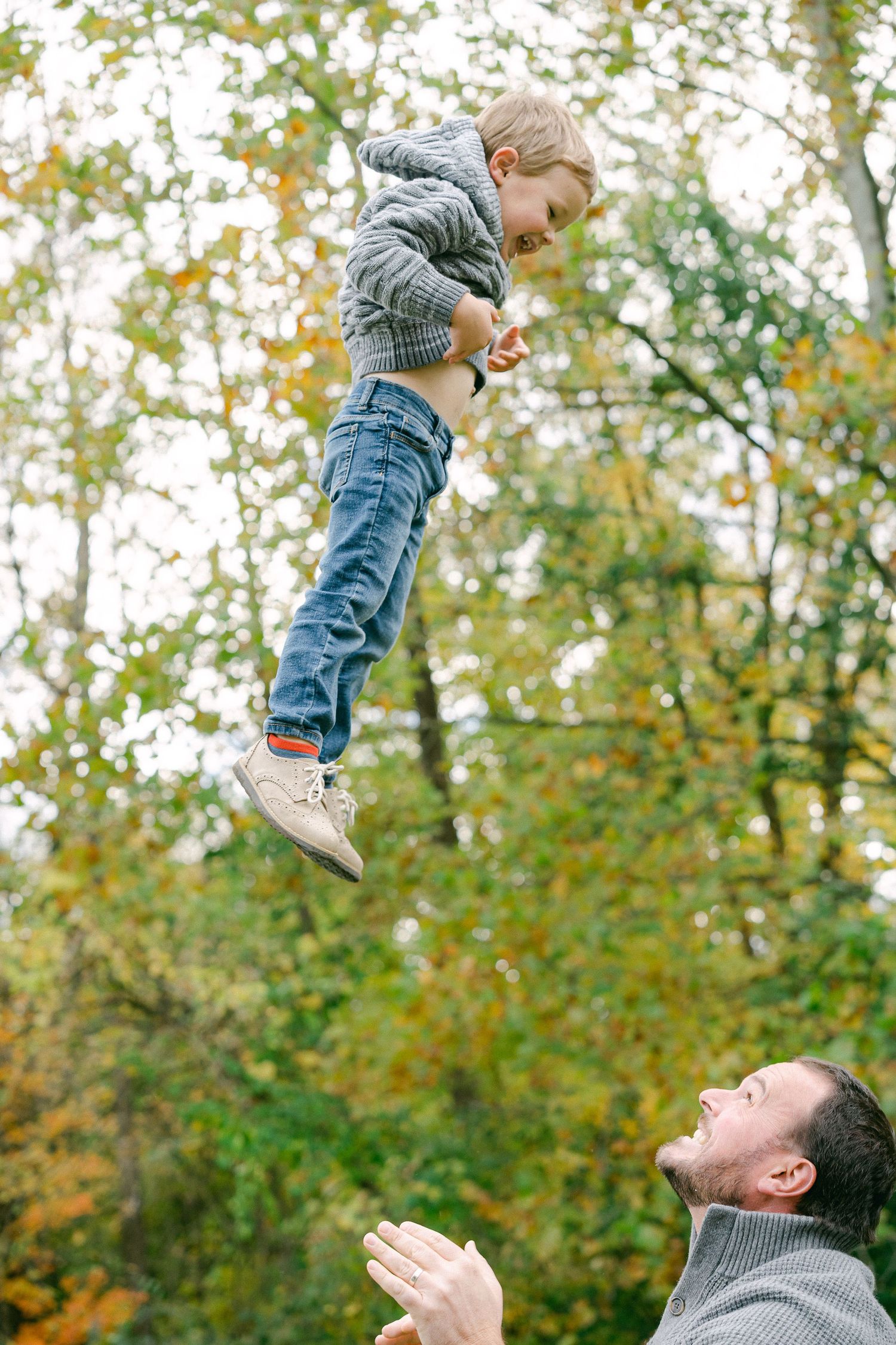 A sequence of playful lifting motions showing a person in grey raising a child up high with trees in the background.
