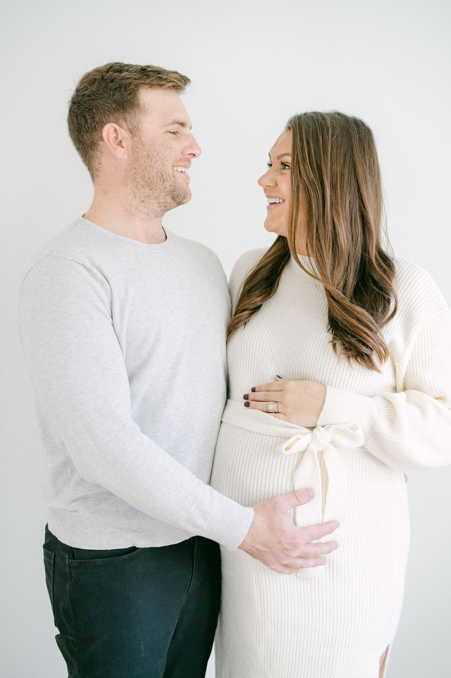 Couple in white sweaters share an intimate moment during maternity photoshoot against light background.