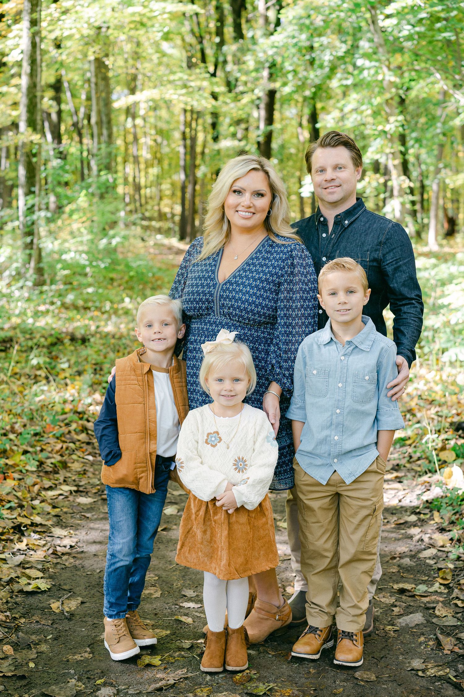 A family poses together on a wooded path during fall with leaves scattered on the ground.