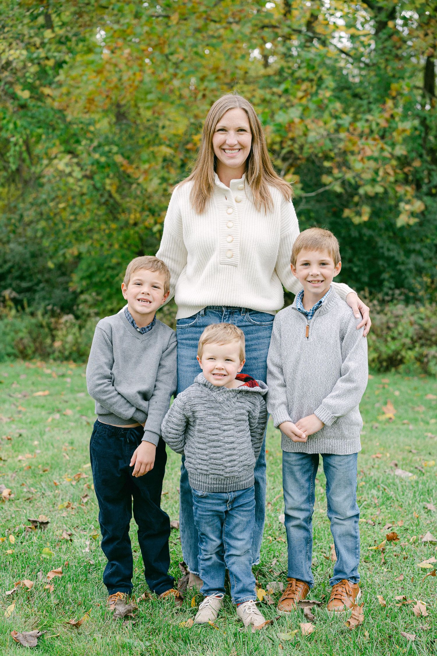 A family group poses together on grass with autumn trees in the background wearing casual sweaters and jeans.
