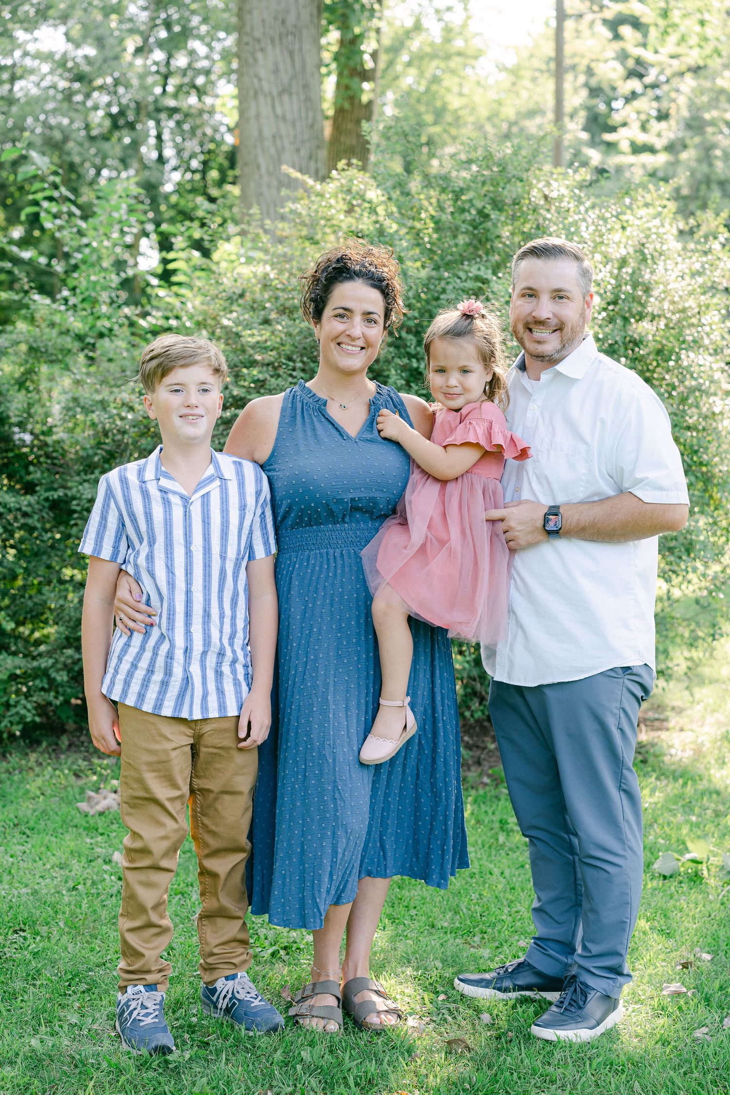 A family stands together outdoors in a park setting with green trees and foliage in the background.